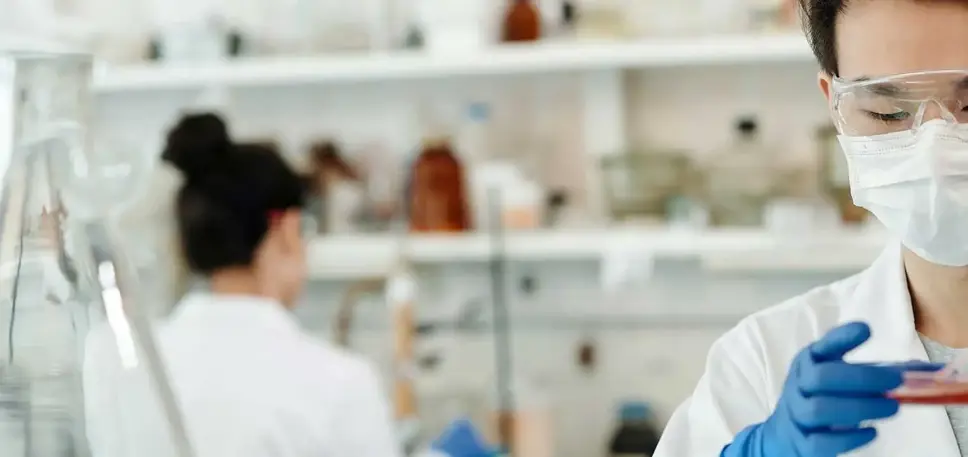 Two scientists in a lab, one the background and the one in the foreground adding material to a petri dish.