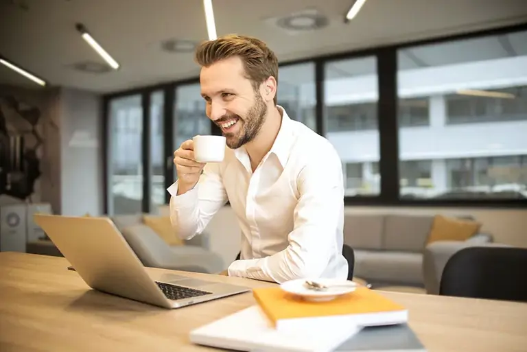 Smiling office worker sits in front of this laptop, about to drink from a cup raised to his lips