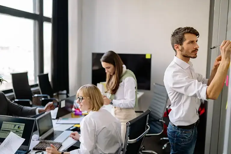 Two team members working at a long office table while another stands and writes a post it note on the wall