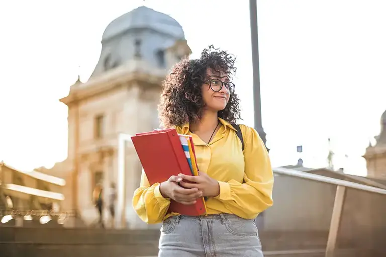 Young woman holding a laptop and notebook in her arms, stands on the steps of a large church, in a setting which looks like an overseas location 