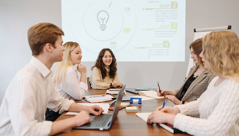 Team of office workers sit at a large table with the presentation projected on the rear wall