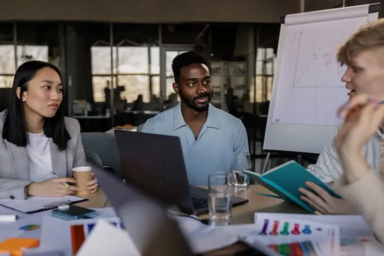 The office workers sit in a office around the table with papers, laptops and whiteboard