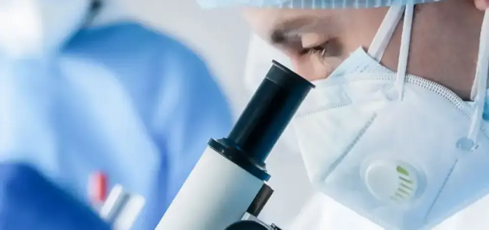 Close up side view of a masked scientist looking down a microscope