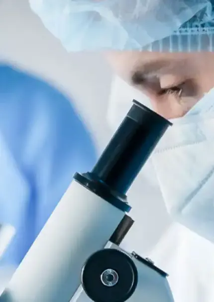 Close up side view of a masked scientist looking down a microscope