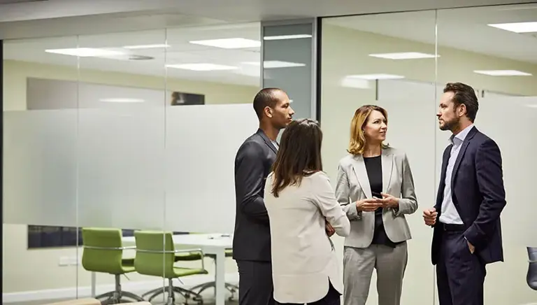 Four office workers stand talking in front of a set of glass meeting rooms