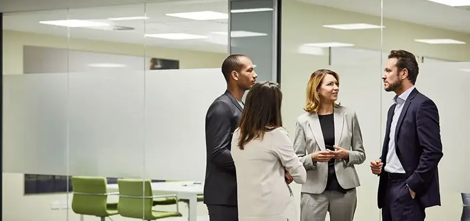 Four office workers stand talking in front of a set of glass meeting rooms