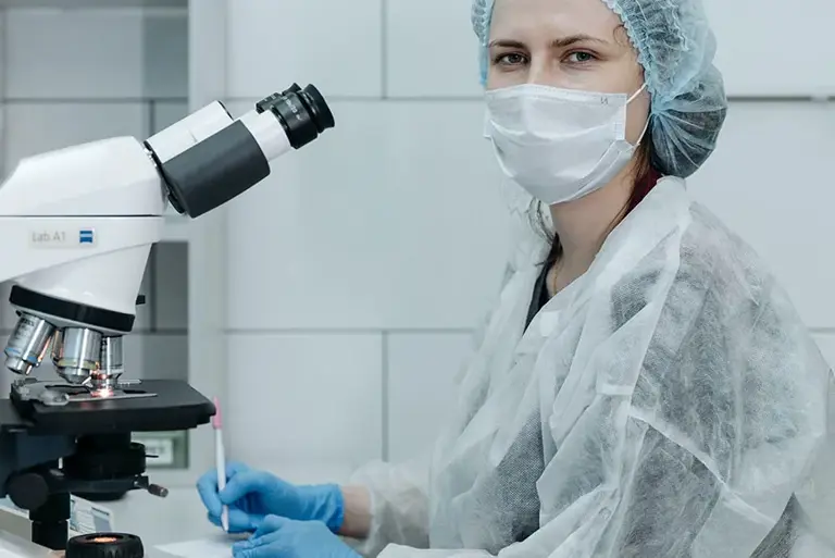 Seated masked scientist, in a lab, looks at us while sat at microscope