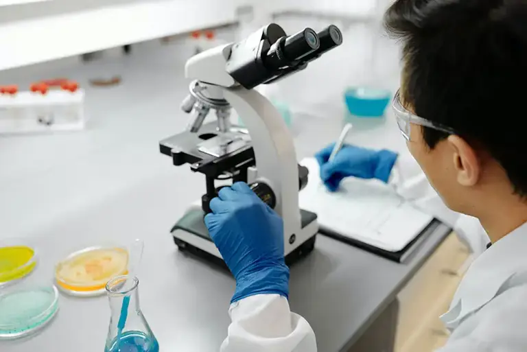 Scientist sits working at a microscope in a lab, writing notes.  