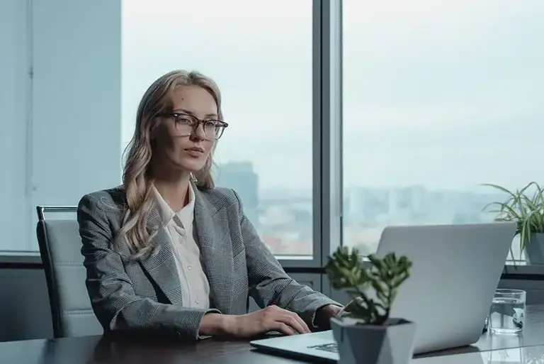 Office worker sits typing at a laptop, at a desk with an impressive city panorama seen through the window behind.