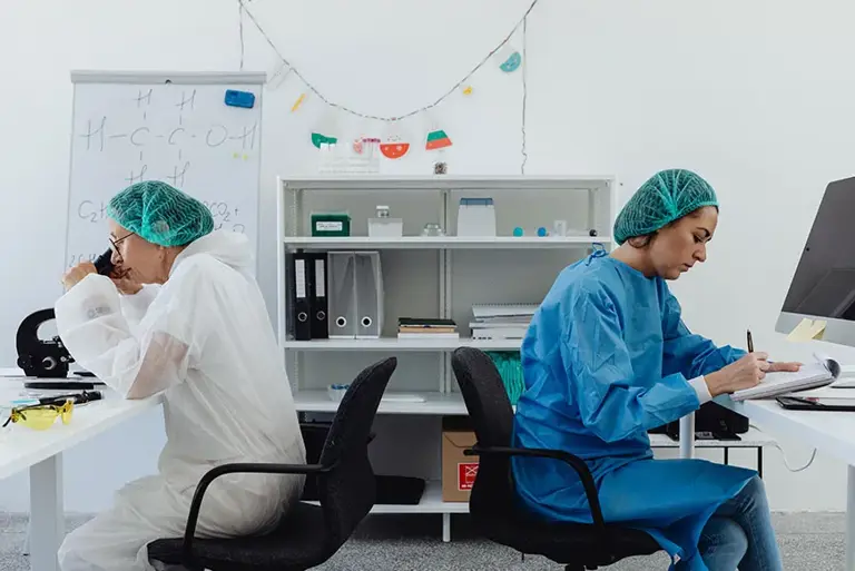 Two scientist sit back-to-back on a lab, one writes notes while the other looks through a microscope