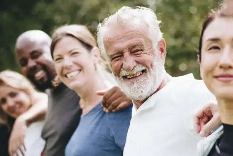 A line of people of all ages and diverse ethnic backgrounds smile to the camera while they all have their arms around each other's backs