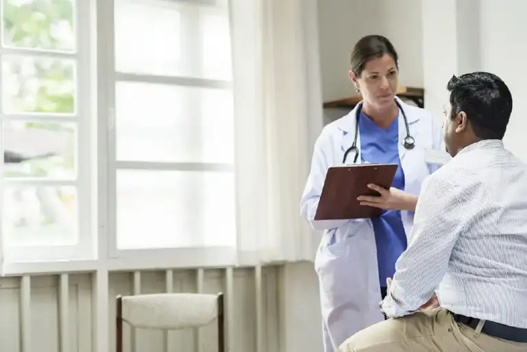 Doctor with a clipboard stands interviewing a patient sitting on a bed in a hospital setting